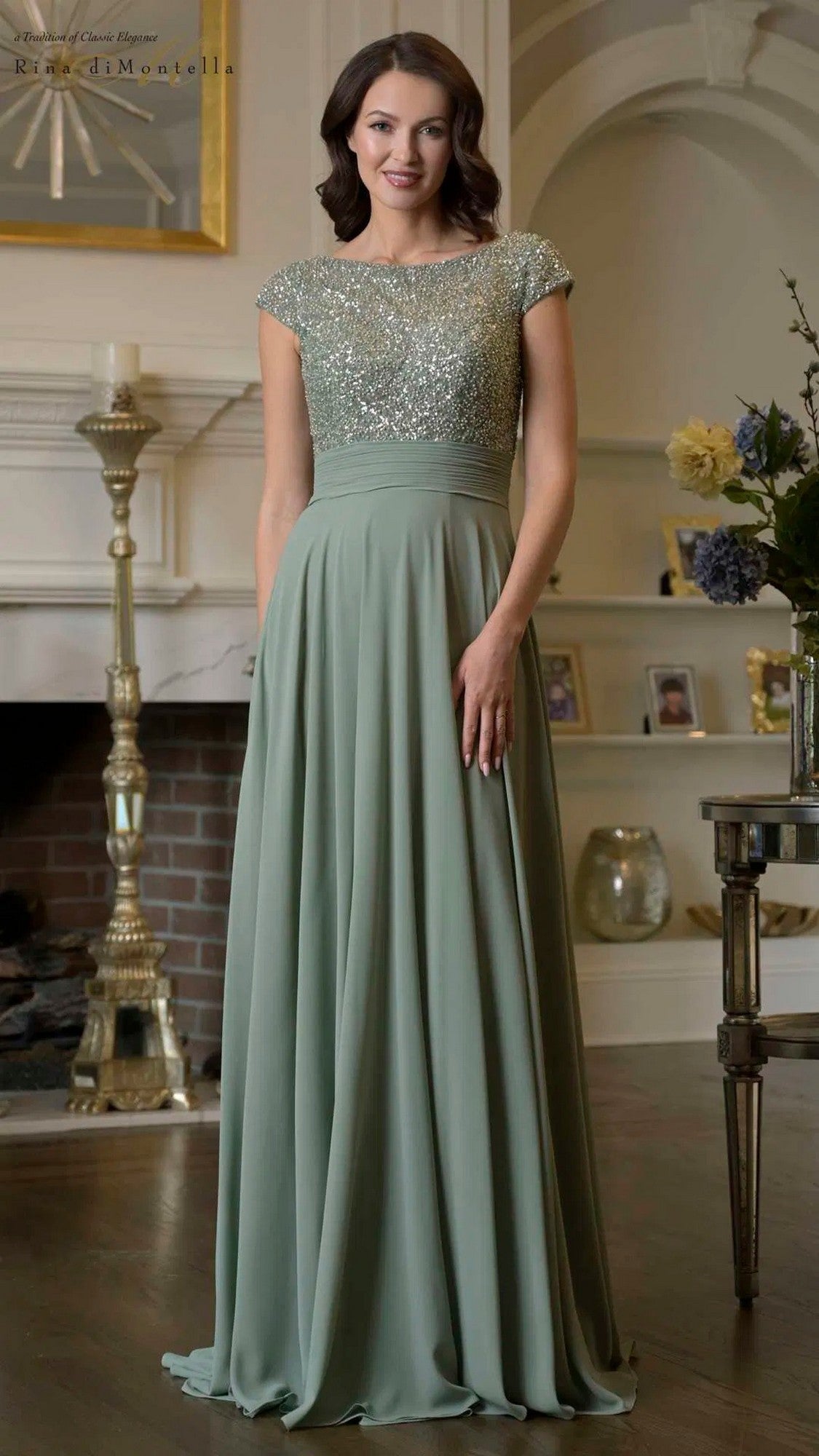 A woman stands indoors wearing the Rina Di Montella RD3419 long formal dress in sage, featuring a sparkling beaded bodice with short sleeves and a floor-length chiffon skirt.